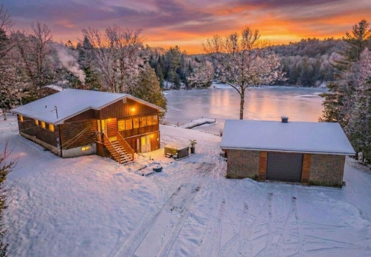 Une cabane au revêtement extérieur en bois, avec un porche abrité, et un paysage enneigé comprenant un lac gelé et des arbres.
