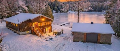 Une cabane au revêtement extérieur en bois, avec un porche abrité, et un paysage enneigé comprenant un lac gelé et des arbres.