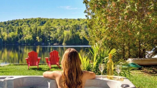 Uma pessoa relaxando em uma banheira de hidromassagem com vista para um lago e uma floresta.