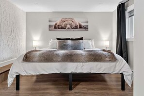 Un dormitorio con una cama grande, dos lámparas de noche, un cuadro colgado en la pared con un oso y una ventana con cortinas.
