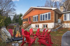 A two-story wooden house with a stone foundation, a wooden deck, and a fire pit with red chairs. - Bécassines | Private Spa | Billiards | Waterfront (Nominingue)