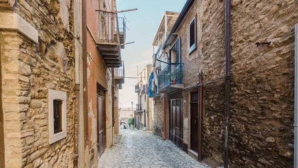 Une ruelle étroite, pavée de pierres, située entre des immeubles dotés de balcons, et fermée par une grille métallique à son extrémité.