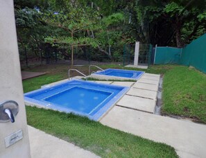 Duas piscinas quadradas com azulejos azuis, rodeadas por relva e árvores, com um terraço de betão em primeiro plano.