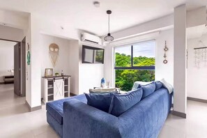 Una sala de estar moderna con un sofá modular, un comedor con mesa y sillas, un ventanal con vista a follaje, y un equipo de aire acondicionado.