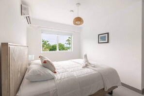 Un dormitorio con cama, ventana, aire acondicionado instalado en la pared y lámpara colgante.