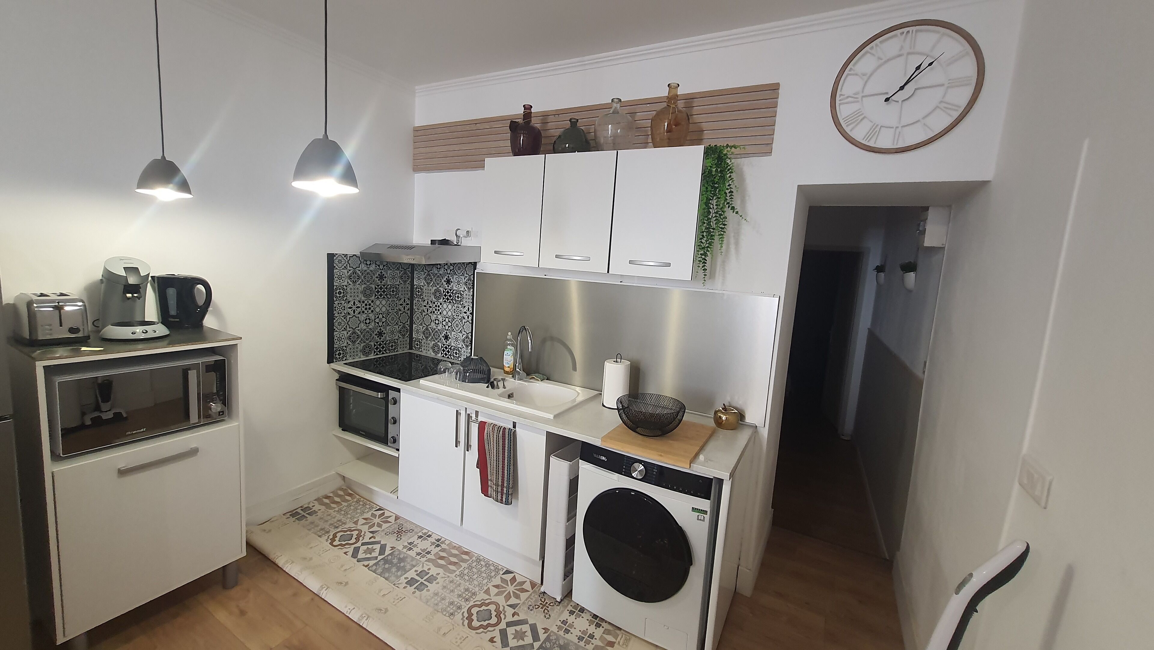 Une cuisine compacte équipée d’un four à micro-ondes, d’un grille-pain, d’une machine à café et d’un évier. Il y a une horloge au mur et un lave-linge dans un coin.