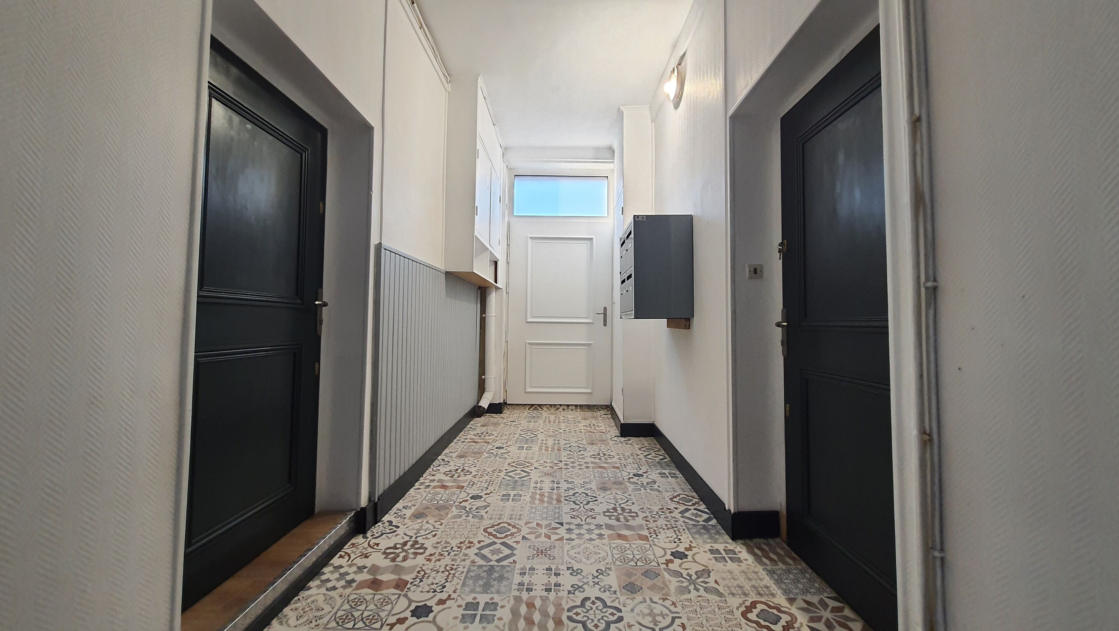 Um corredor estreito com ladrilhos estampados, portas pretas em ambos os lados e uma porta branca no final.