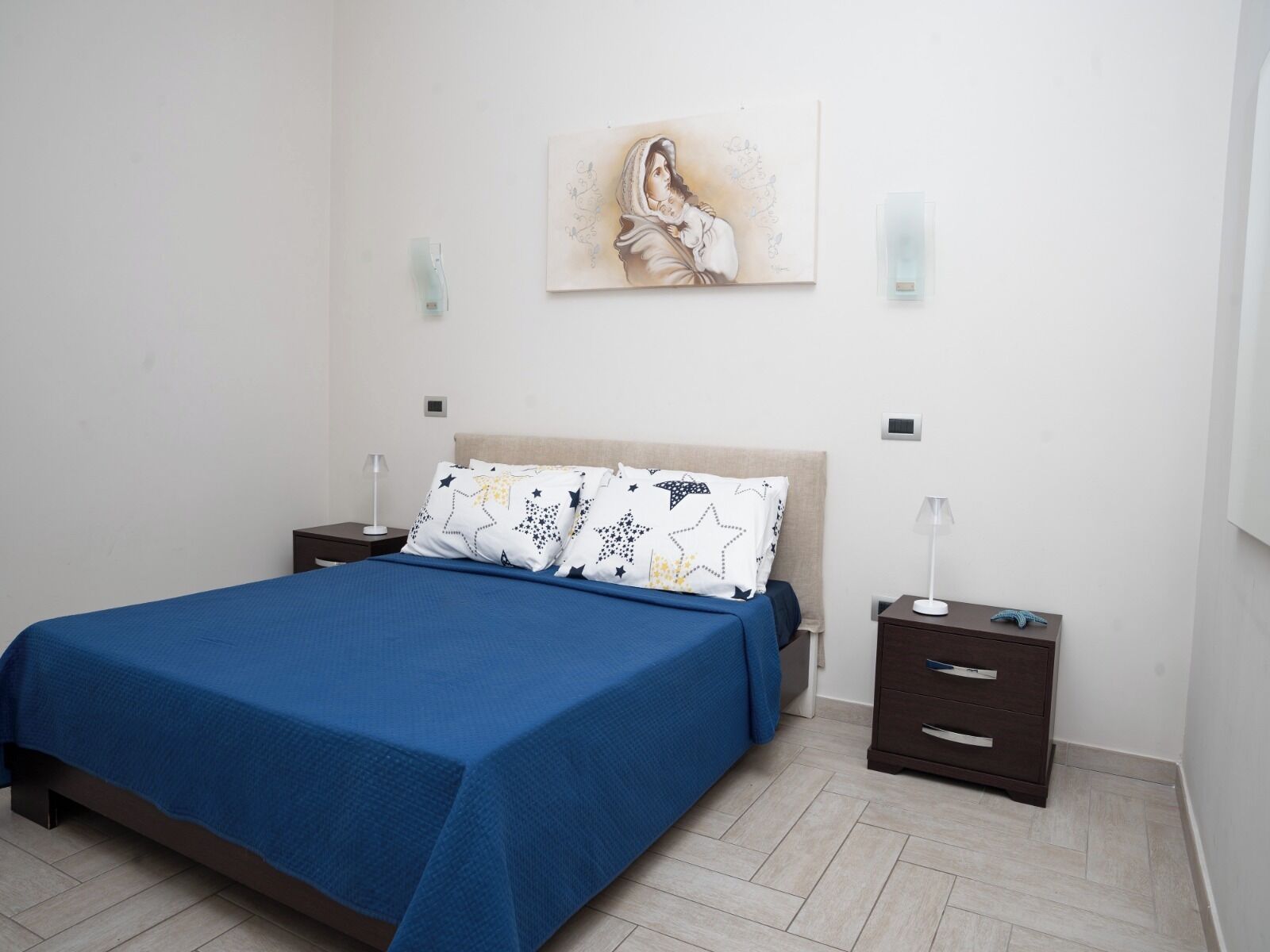 Une chambre à coucher comprenant un lit, deux tables de chevet, une commode, une lampe fixée au mur et un tableau.