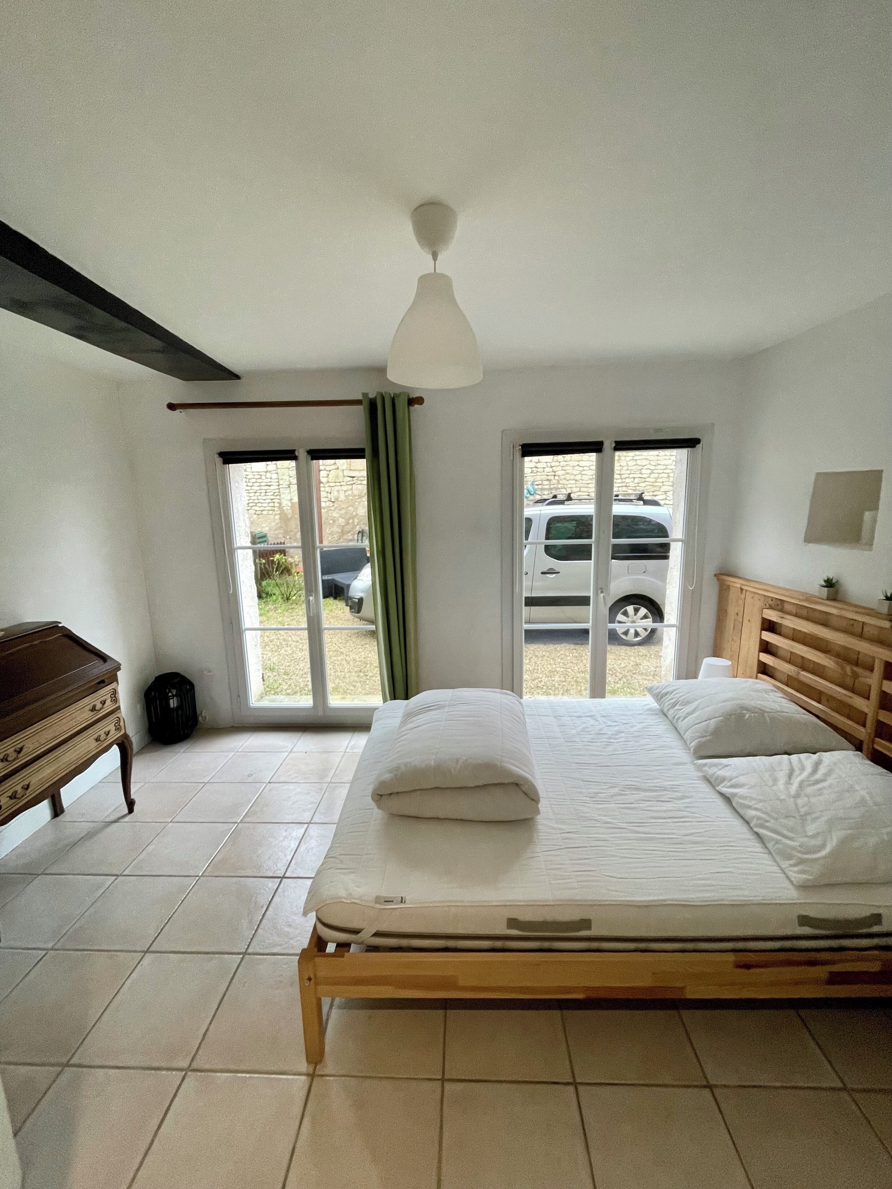 A bedroom with a bed, a wooden dresser, a ceiling fan, and a view of a parked car through the window.