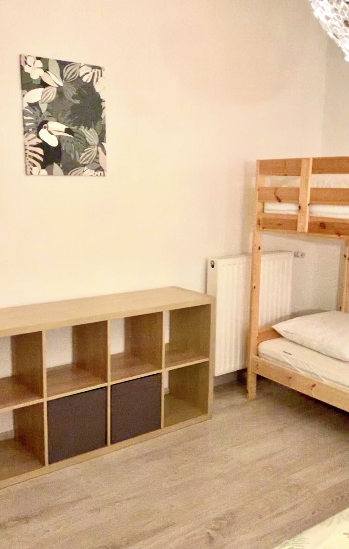 A bunk bed with a wooden frame and a lower shelf, a radiator, and a wall-mounted picture.