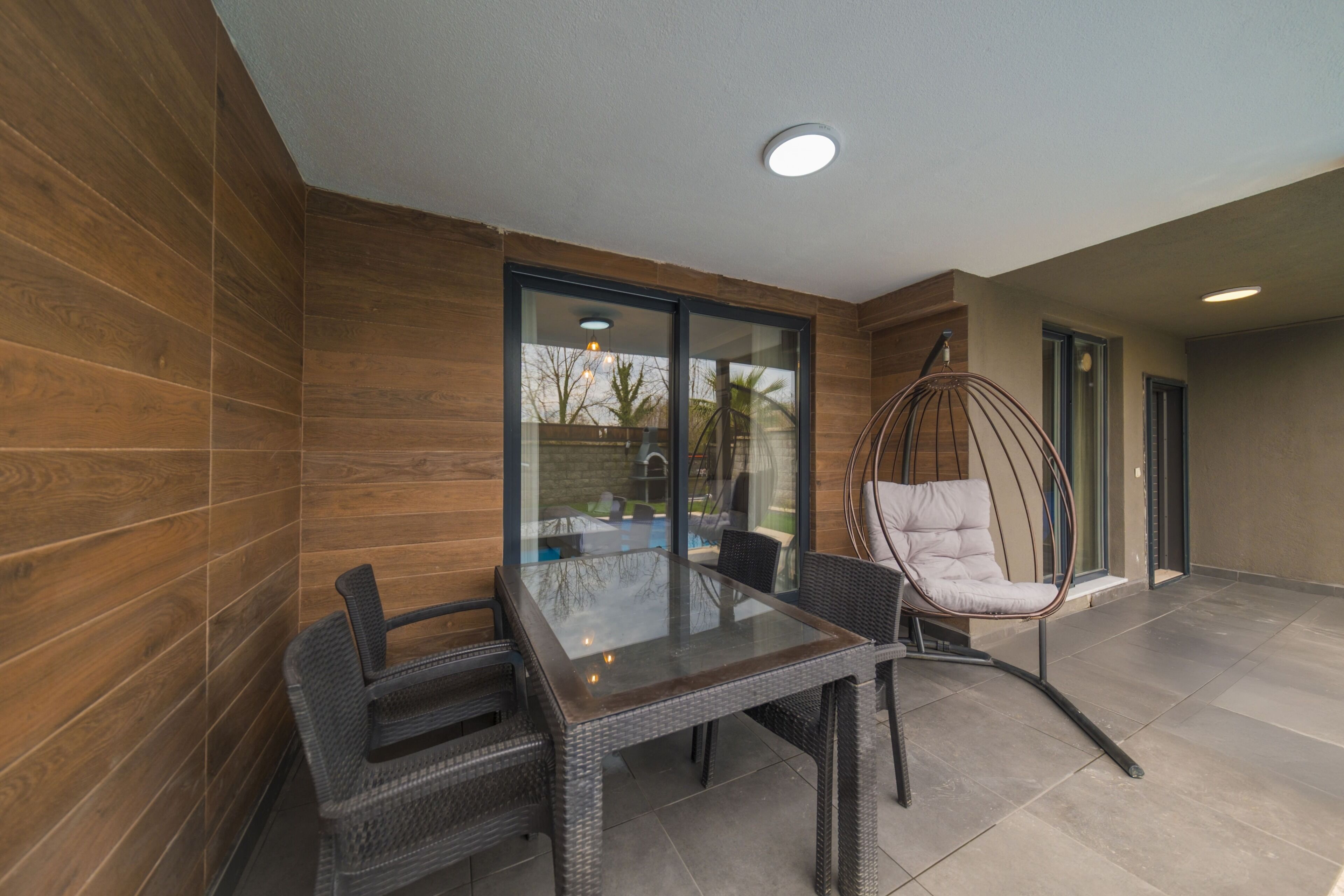 A patio with a glass table and wicker chairs, a hanging chair, and a view of the outside.