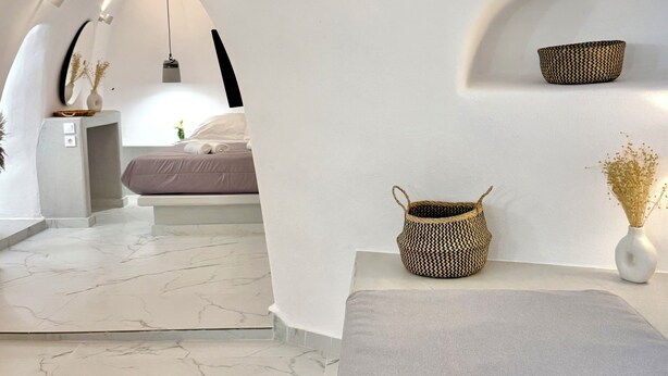 A modern interior with a marble floor, a bed with a white headboard, a small table with a vase, and a hanging light fixture. - Petuna Cave Suites (Santorini)