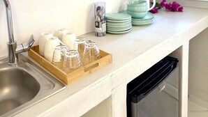 A kitchen with a stainless steel sink, a wooden tray with glassware, and stacked plates and cups on the counter. - Petuna Cave Suites (Santorini)