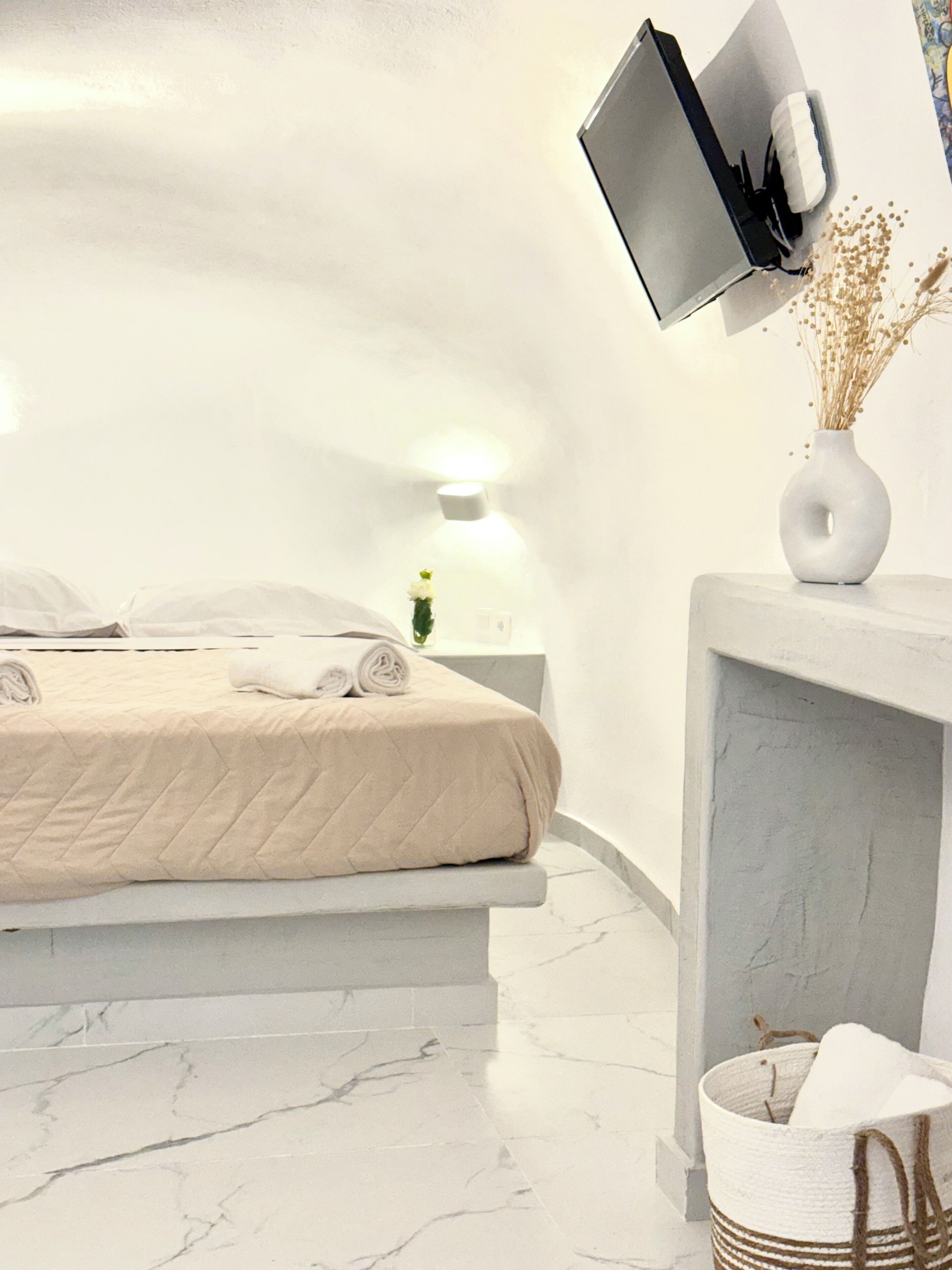 A modern bedroom with a bed, a TV mounted on the wall, a bedside table with a lamp, and a vase with dried flowers.