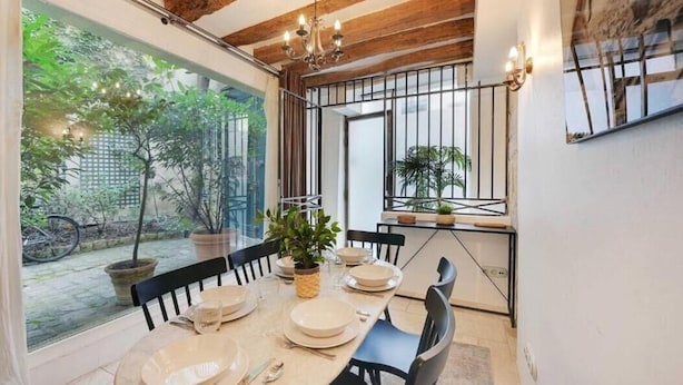 A dining area with a table set for four, a chandelier, and a view of a garden through large windows. - Family Apartment - Quartier Latin (Paris)