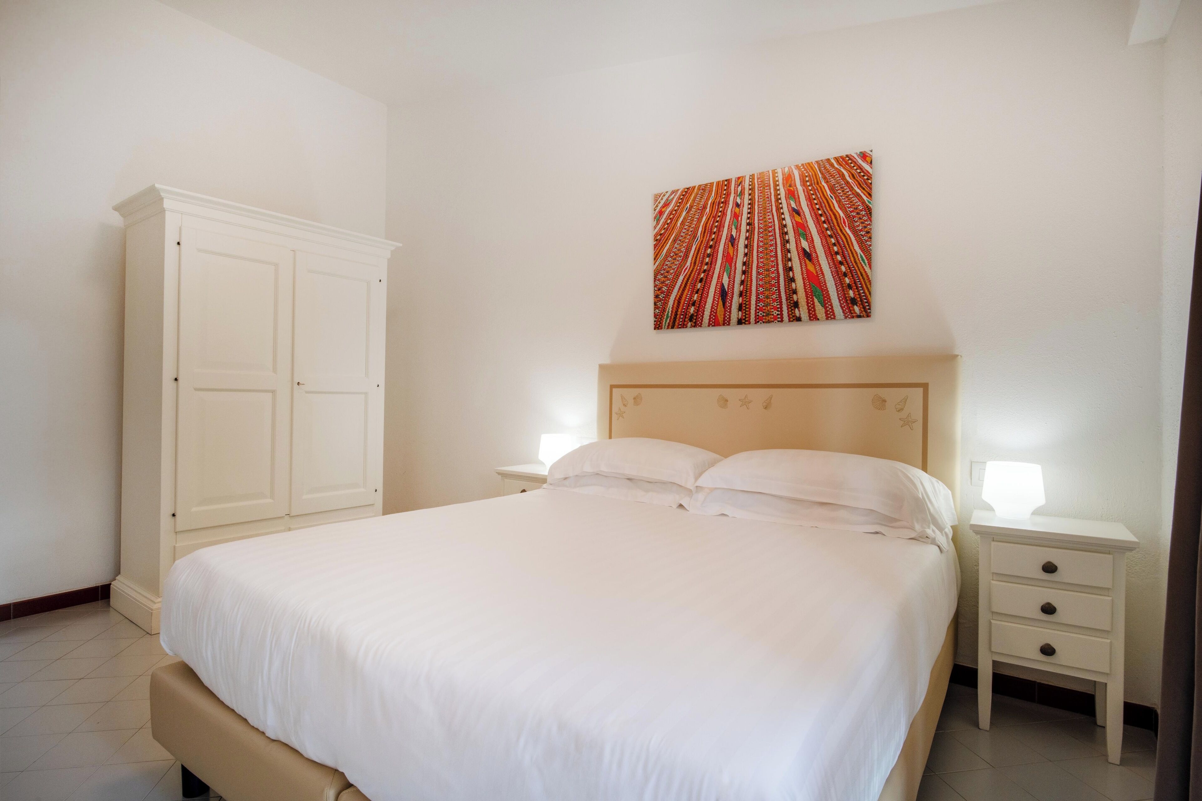 Une chambre bien rangée, avec un grand lit, une armoire, une table de chevet avec une lampe et une œuvre d’art colorée accrochée au mur.