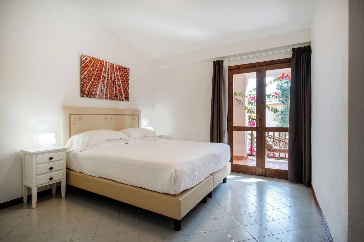 A bedroom with a bed, a nightstand, a wall art piece, and a view of the outdoors through a door.