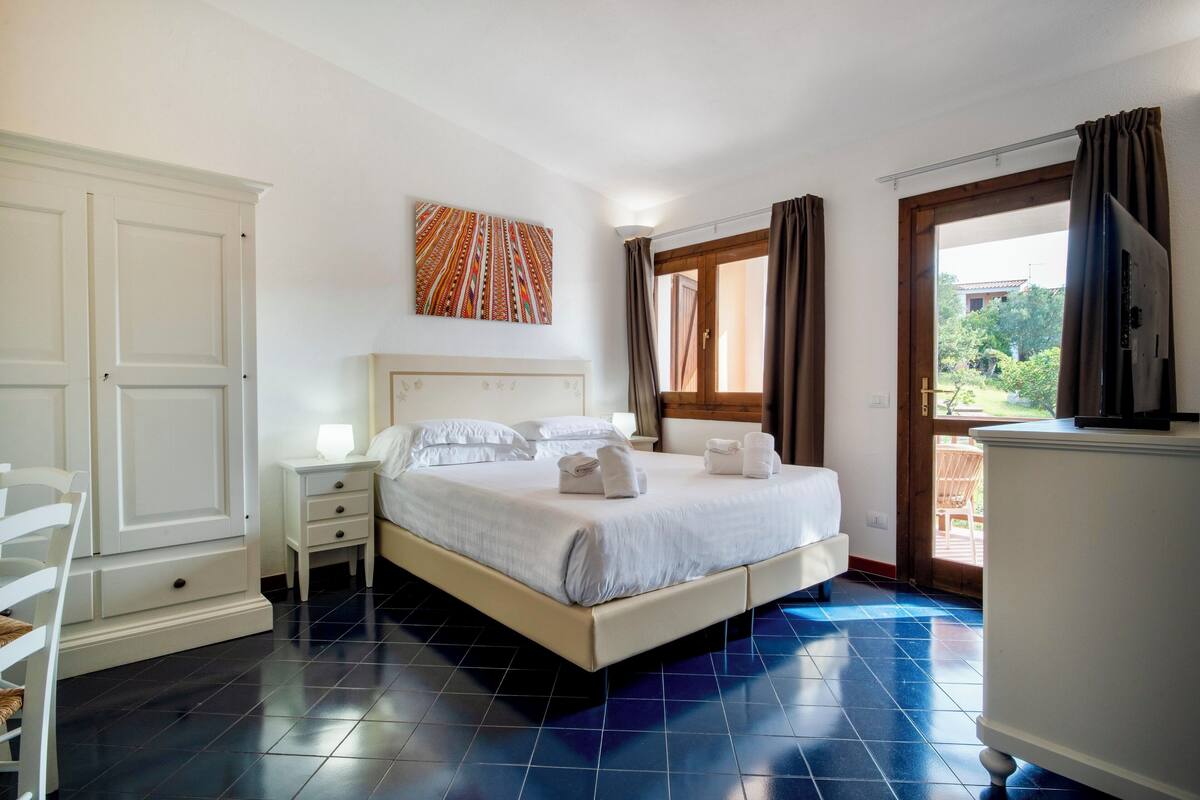 A hotel room with a bed, bedside tables, a wardrobe, a window with curtains, and a view of the outdoors.