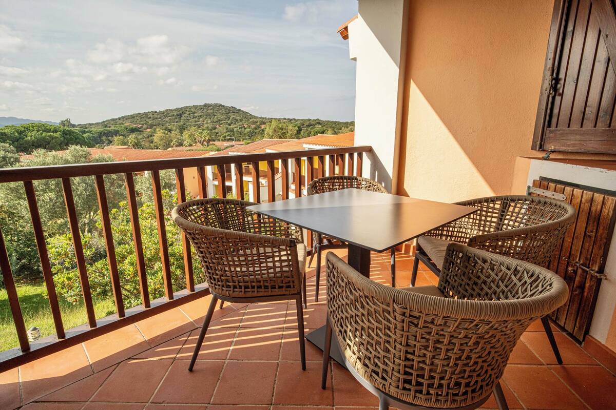 A balcony with a table and wicker chairs, overlooking a scenic landscape with hills and trees.