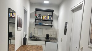 Um corredor estreito com uma área de cozinha, incluindo bancada, pia e fogão. Há um banquinho branco pequeno e uma porta à direita.