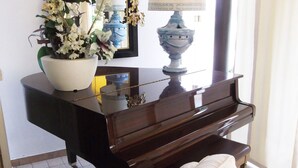 Ein Klavier mit weißem Kissen, einer Blumenvase und einer blauen Lampe auf einem dunklen Holzständer in einem Raum mit Fliesenboden.