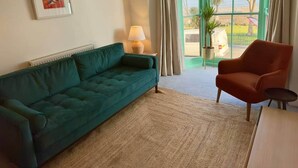 Una sala de estar con un sofá verde, un sillón rojo, una lámpara, un cuadro enmarcado y vistas al exterior por una ventana.