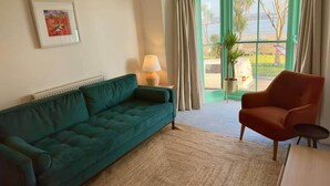 Una sala de estar con un sofá verde, un sillón rojo, una lámpara, un cuadro enmarcado y vistas al exterior por una ventana.