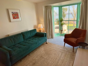 Una sala de estar con un sofá verde, un sillón rojo, una lámpara, un cuadro enmarcado y vistas al exterior por una ventana.