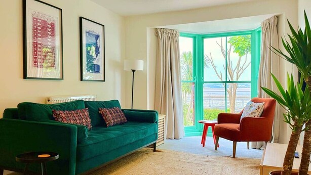 Una sala de estar con un sofá verde, una silla roja, una planta en maceta y un ventanal con cortinas.