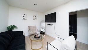 A modern living room with a black sofa, a white armchair, a glass coffee table, and a TV mounted on the wall. - Cozy East Downtown Houston Getaway Near UH, Med Center, NRG Stadium (Houston)
