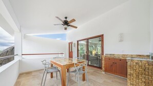 En balkon med et bord og stole, en ventilator i loftet, en skydedør af glas og et tekøkken med vask og bordplade.