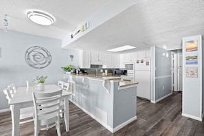 A modern kitchen with white cabinetry, a central island, and a dining area with white chairs. - Waterpointe II 704 - Spacious 3 Bedroom Condo (North Myrtle Beach)