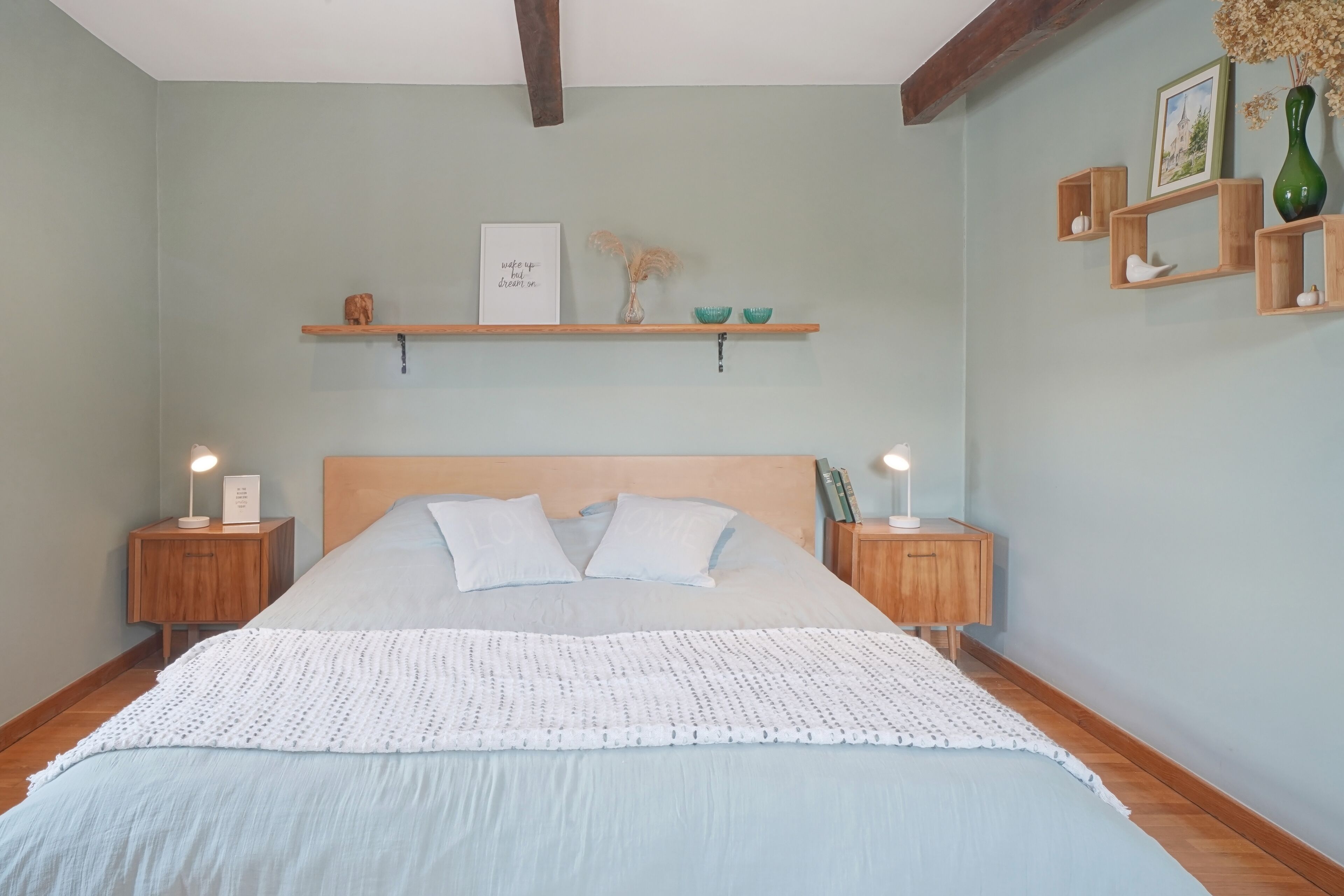 A bedroom with a large bed, wooden headboard, two bedside tables with lamps, a wall-mounted shelf with decorative items, and a framed picture.