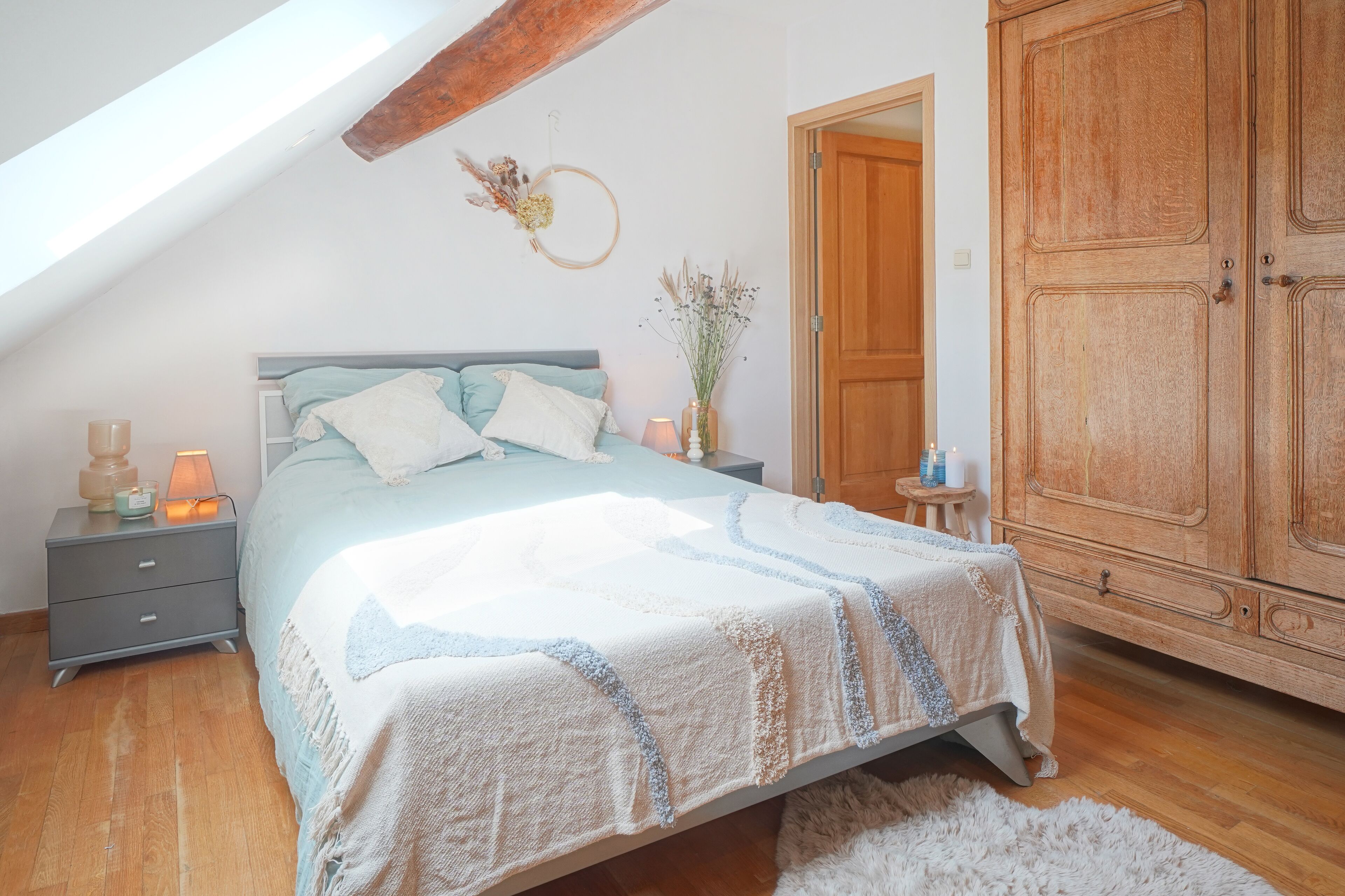 A bedroom with a sloped ceiling, a bed with a light blue and white bedspread, a wooden wardrobe, and a small bedside table with a lamp.