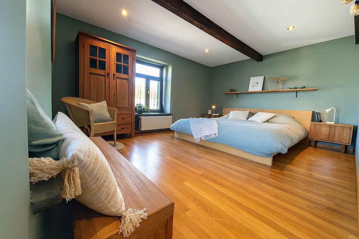 Un dormitorio con una cama de madera, un armario de madera, una silla de mimbre, una mesita de noche con una lámpara y una ventana con vistas al exterior.