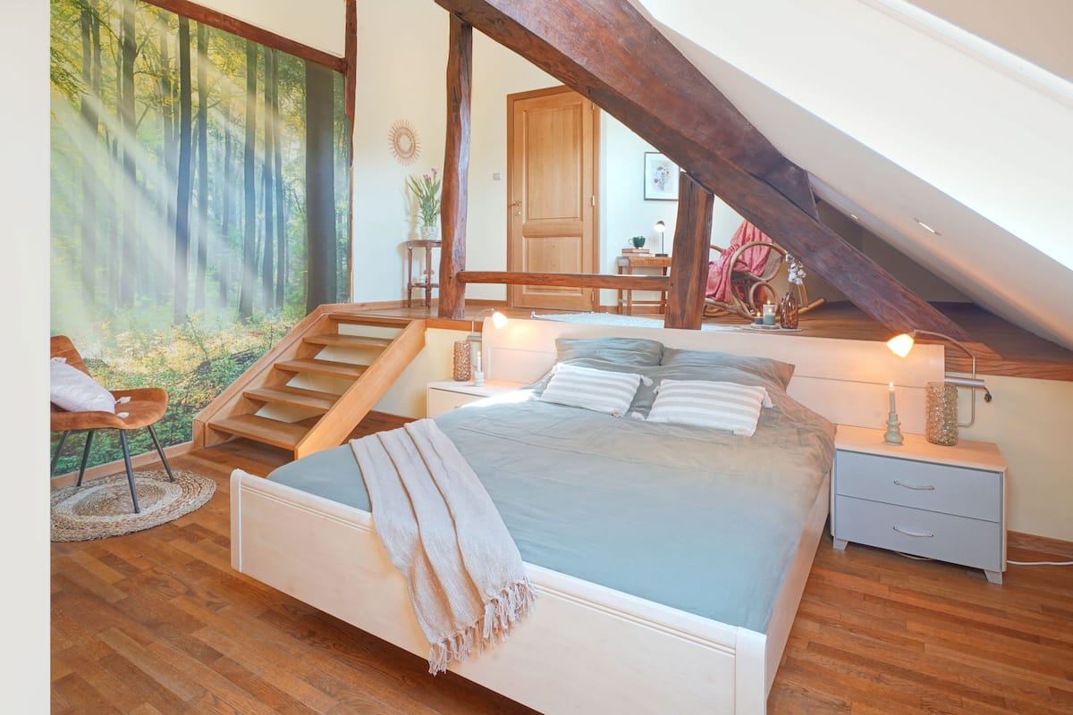 Un dormitorio con una cama grande, vigas de madera, vista a un bosque por una ventana y una escalera que sube a un nivel superior.