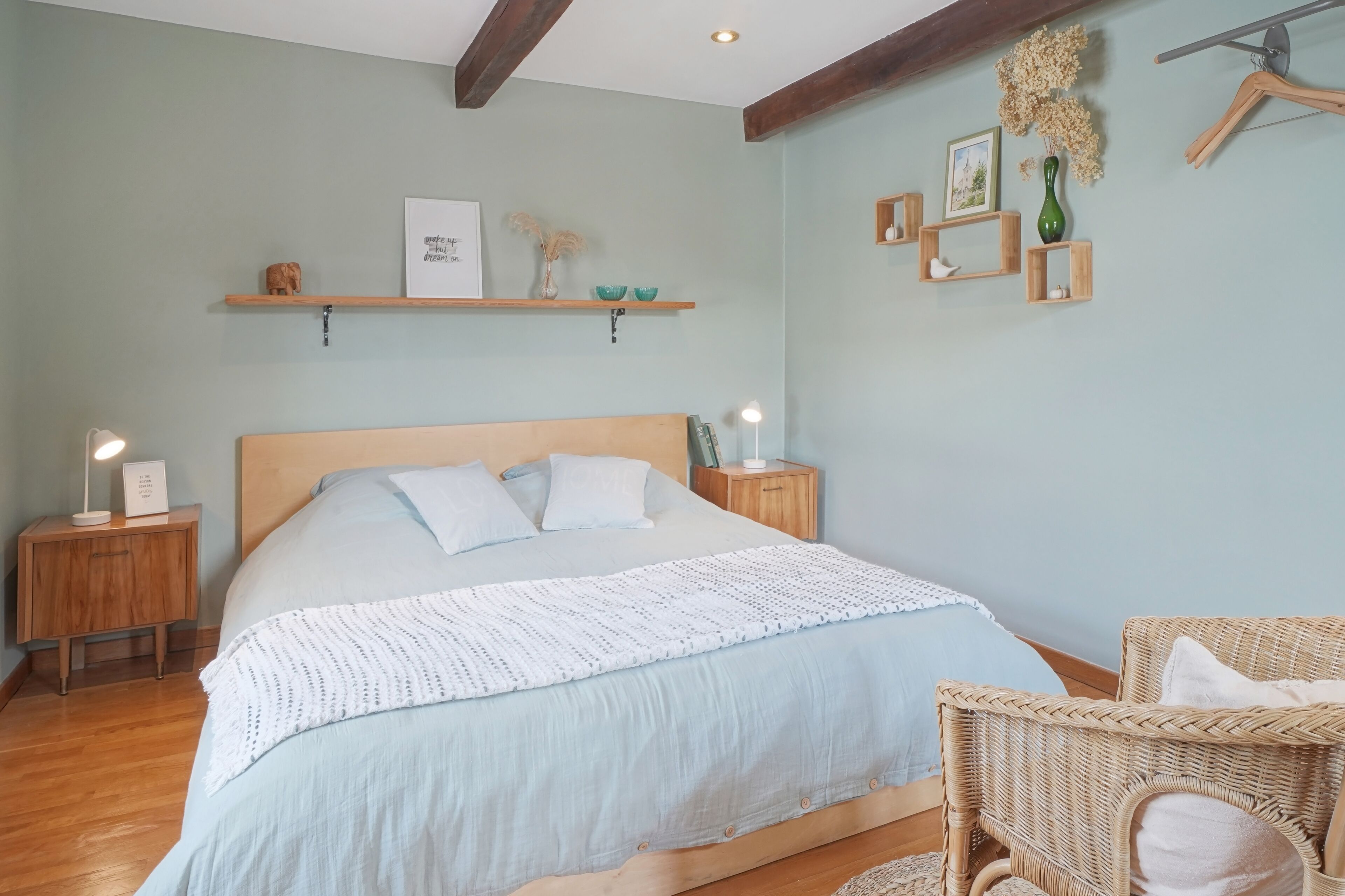 A bedroom with a large bed, wooden nightstands, a wicker chair, and wall-mounted shelves with decorative items.