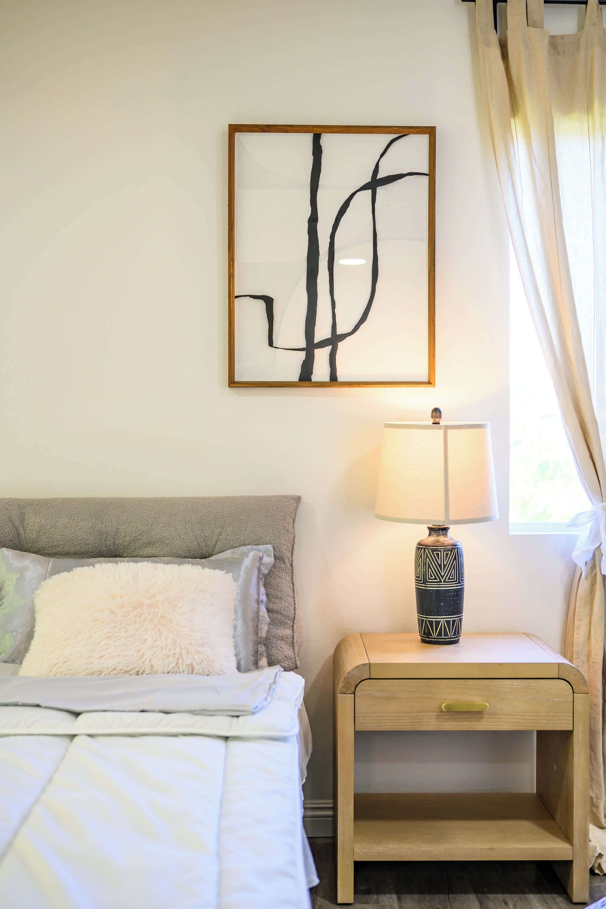 Een slaapkamer met een bed, een nachtkastje met een lamp, een kunstwerk met een abstracte lijst, en een raam met gordijnen.