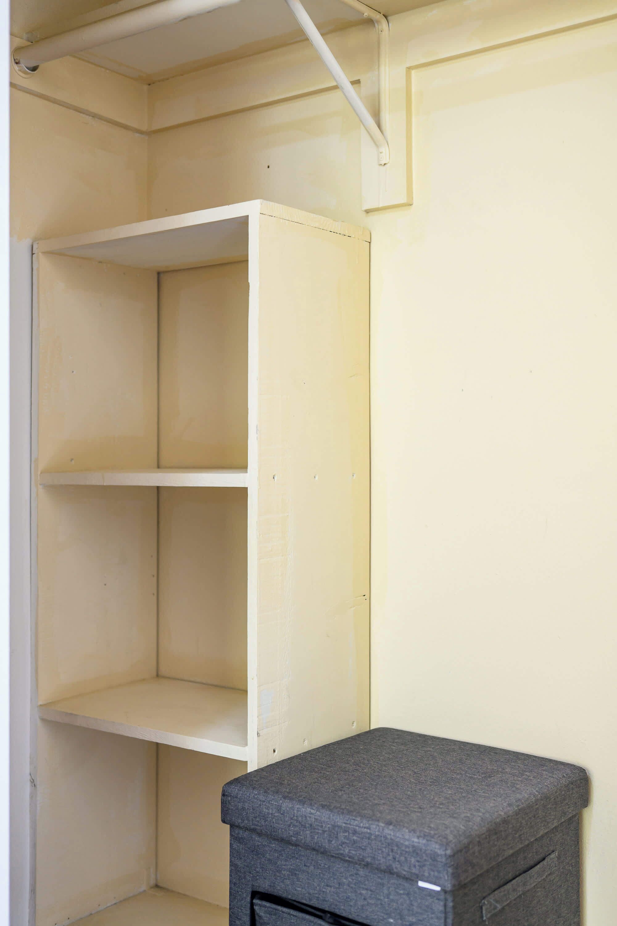 Ein Zimmer mit einer weißen Regaleinheit an einer cremefarbenen Wand, einer grauen Aufbewahrungsbox und einer weißen Rohrleitung an der Decke.