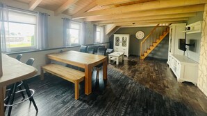A spacious room with a high ceiling, wooden beams, a dining area with a table and chairs, a staircase, and a white cabinet. - Allgäunest Apartment with Mountain View, Private Terrace and Wi-Fi (Argenbühl)