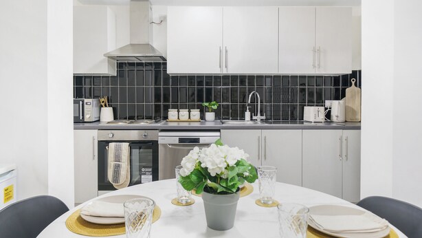 A modern kitchen with white cabinets, a black backsplash, and a dining area with a table set for two. - Shirland Road Flat (London)