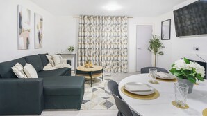 A modern living room with a dark green sofa, a round dining table set with plates and glasses, and a patterned curtain in the background. - Shirland Road Flat (London)
