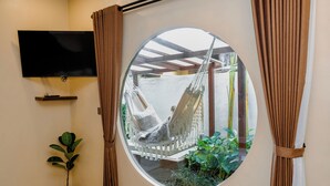 A room with a round window, a TV mounted on the wall, a small potted plant, and a hammock chair. - Lux Villa With Outdoor Pool & Garden (Bali)