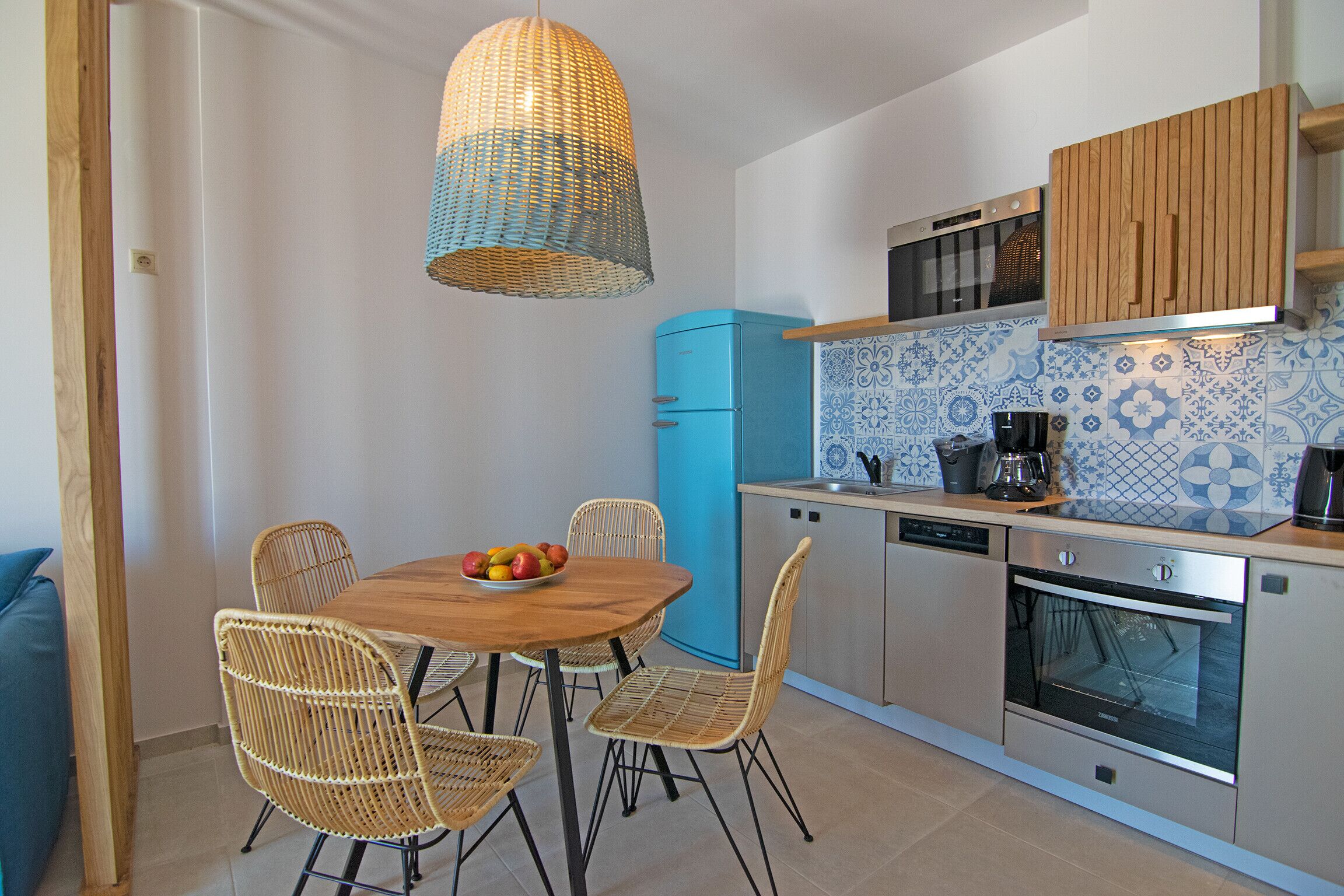 A kitchen with a dining table, chairs, a fruit bowl, a blue refrigerator, and a hanging light fixture.