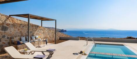 An outdoor pool area with lounge chairs, a small table, and a pergola overlooking the sea.