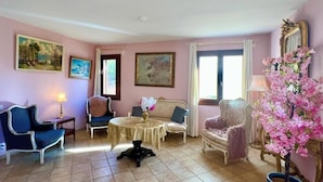 Ένα άνετο σαλόνι με ένα στρογγυλό τραπέζι, δύο καναπέδες και μια πολυθρόνα. Στους τοίχους υπάρχουν πίνακες και σε ένα μικρό τραπεζάκι βρίσκεται ένα βάζο με λουλούδια.