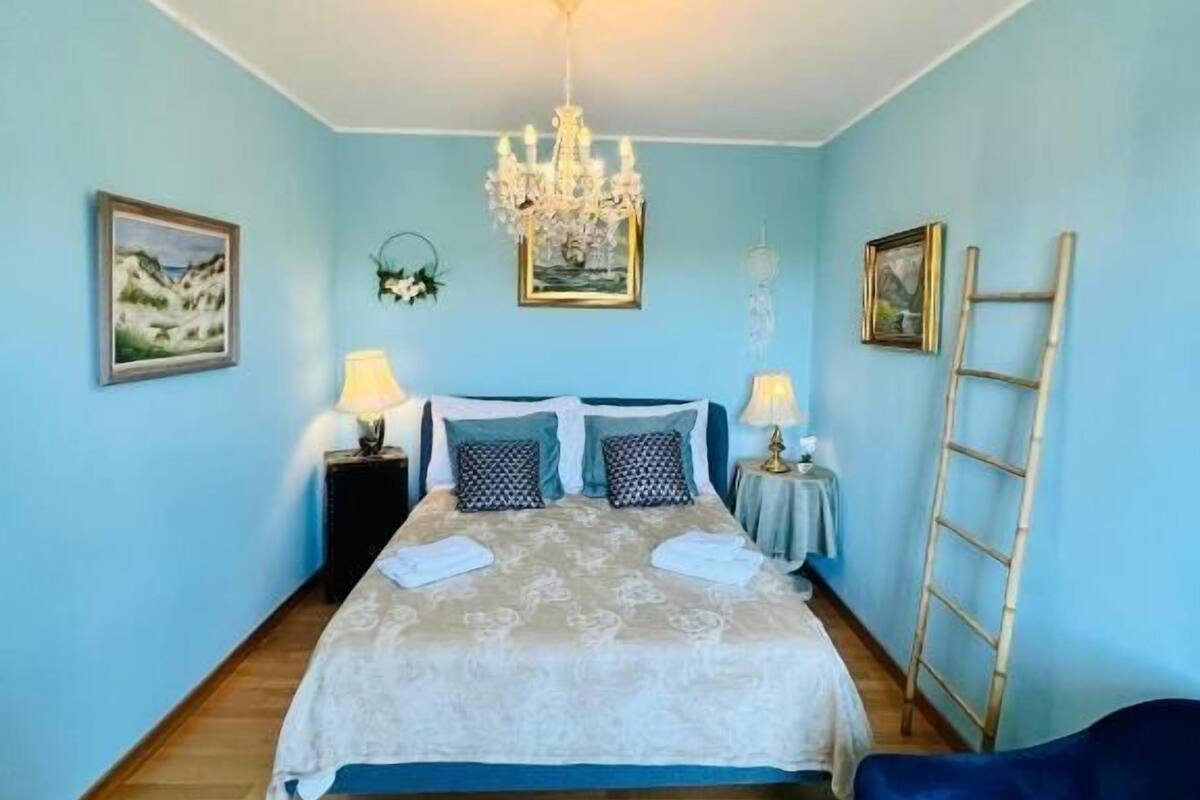 A bedroom with a bed, two bedside tables, a chandelier, and a ladder against the wall.