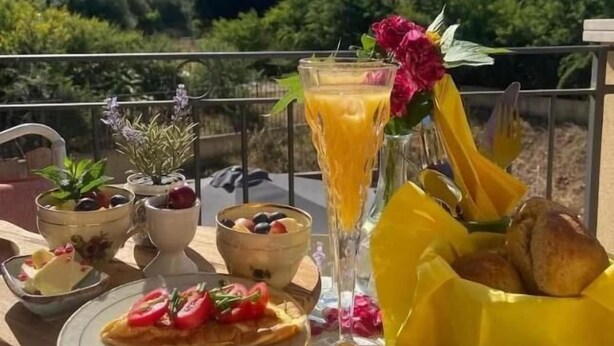 Ένα τραπέζι πρωινού με διάφορα πιάτα, ένα ποτήρι χυμό πορτοκάλι και ένα καλάθι με ψωμί, τοποθετημένα σε ένα γραφικό σημείο με φόντο ένα εντυπωσιακό βουνό.