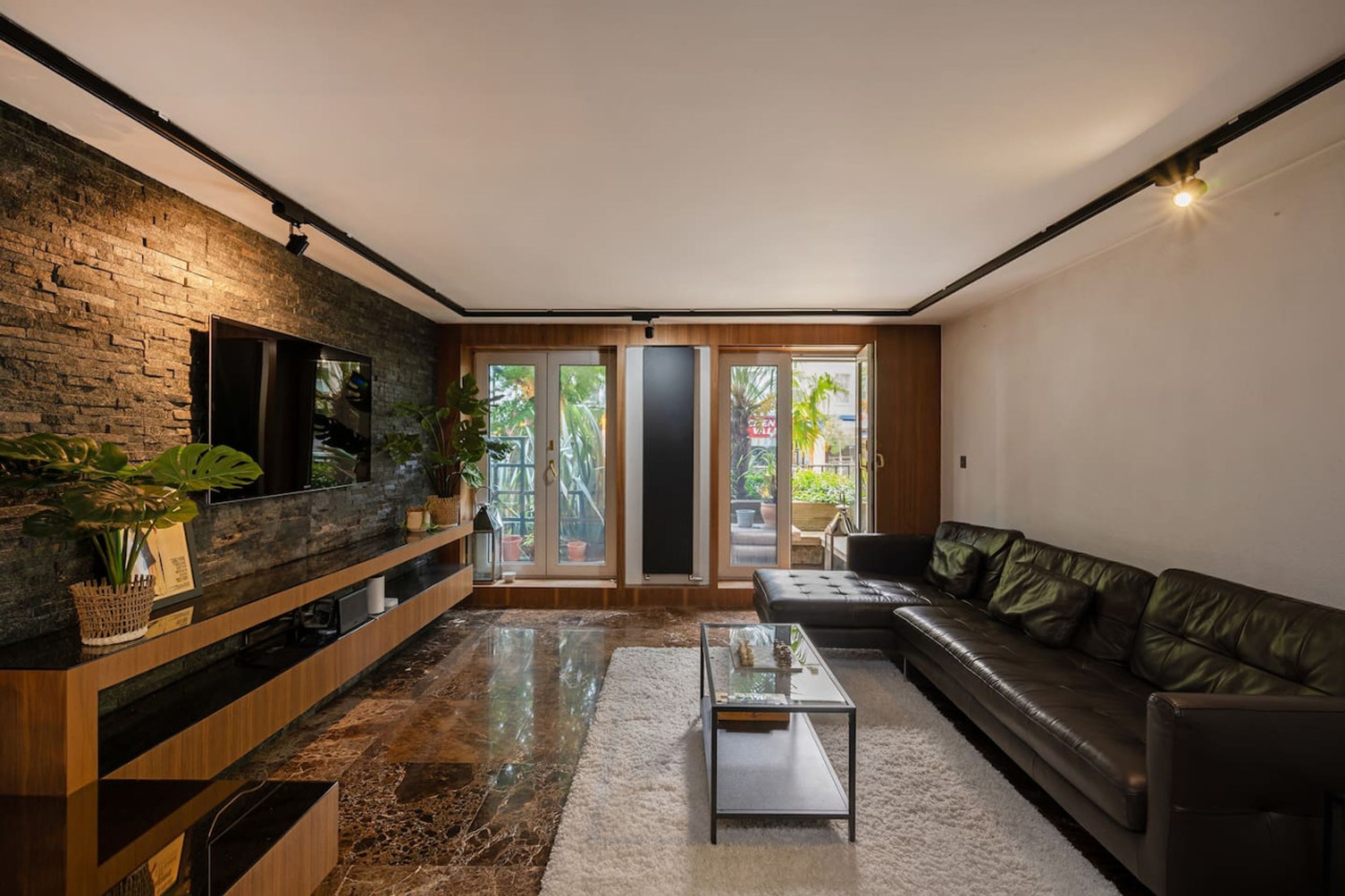 Una sala de estar moderna con una pared de piedra, un sofá de cuero, una mesa de centro de vidrio y un televisor montado en la pared.