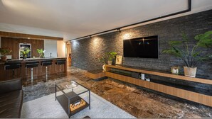 Uma sala de estar moderna com parede de detalhes em pedra, uma televisão de tela plana, uma área de cozinha com armários de madeira e uma mesa de centro.